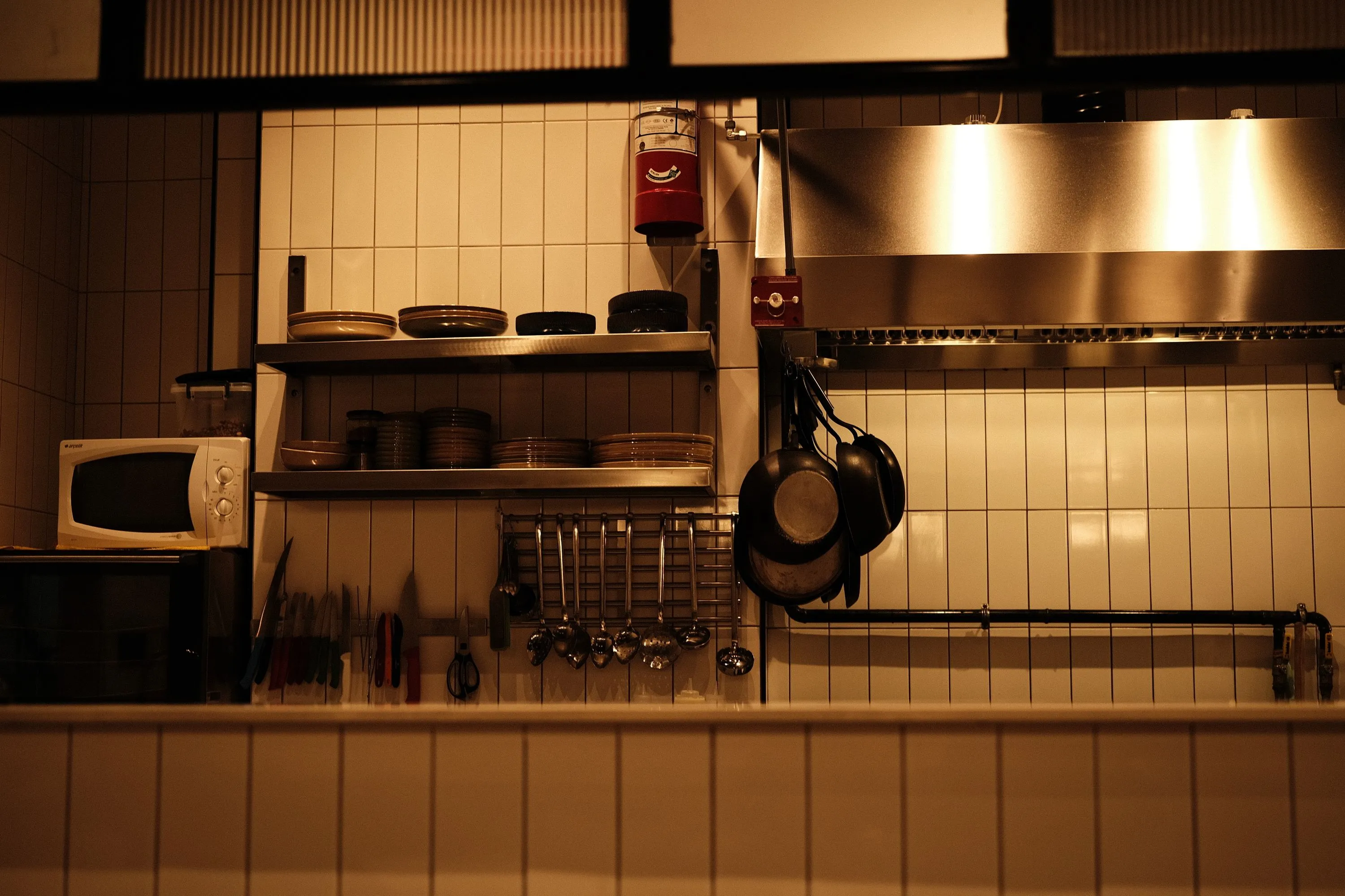 Restaurantkøkken med hylder med tallerkener, ophængte pander og knive på en flisevæg